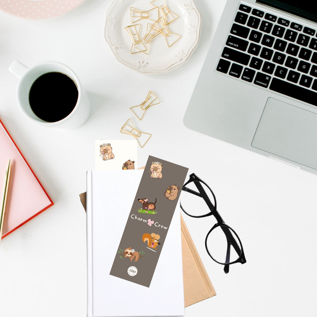 Charm Crew double-sided cardstock bookmark tucked into a notebook on a desk with coffee, laptop, and stationery accessories.