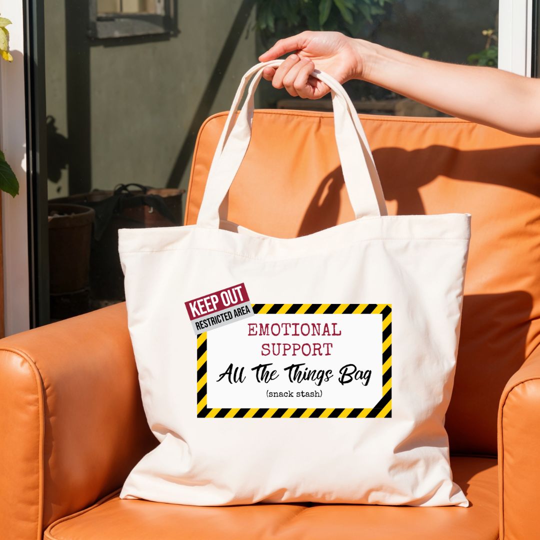 Canvas tote bag with “Emotional Support All The Things Bag” design held in hand on outdoor chair