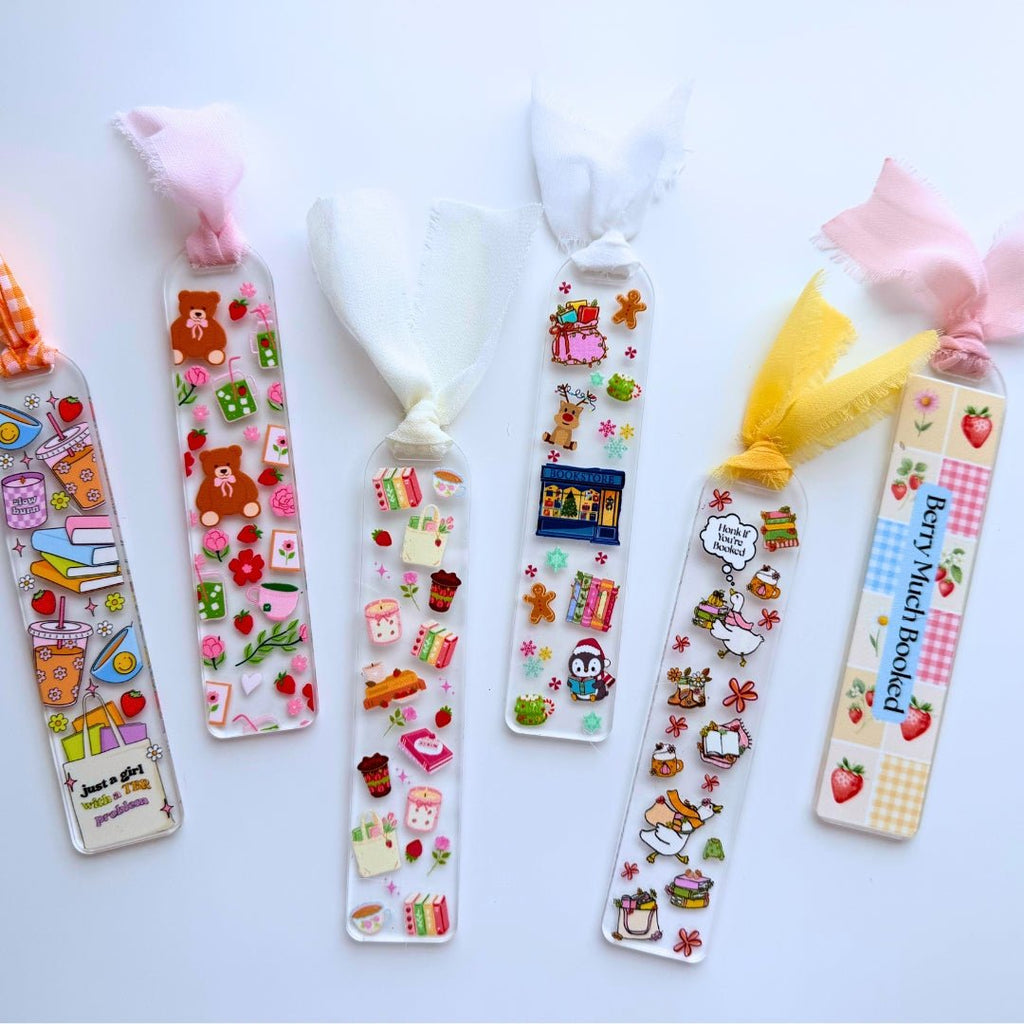 Six clear acrylic bookmarks with colorful bookish designs, including teddy bears, strawberries, books, a goose, and holiday illustrations, finished with coordinating fabric ribbons.