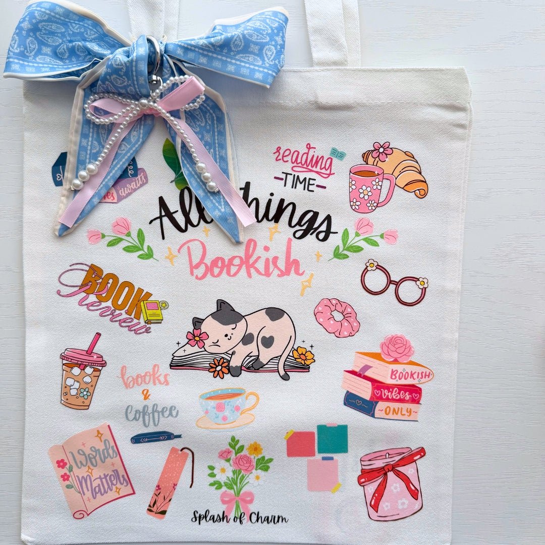 A white canvas tote bag decorated with a blue paisley ribbon and pearl beads, featuring a collection of colorful book-themed illustrations and text including 'All things Bookish' and 'Splash of Charm'.