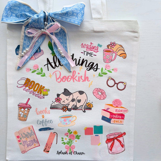 A white canvas tote bag decorated with a blue paisley ribbon and pearl beads, featuring a collection of colorful book-themed illustrations and text including 'All things Bookish' and 'Splash of Charm'.