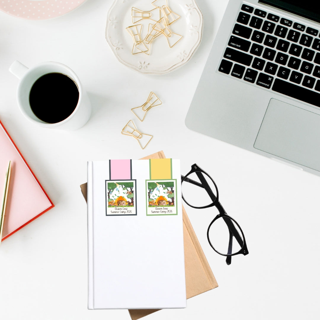 Charm Crew Summer Camp laminated magnetic bookmarks clipped onto a notebook on a desk with coffee, laptop, and stationery accessories.
