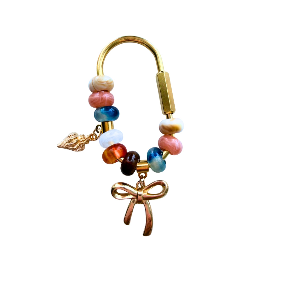 Handmade coastal beaded bag charm with gold bow charm, shell charm, colorful beads, and gold carabiner clasp on a white background.