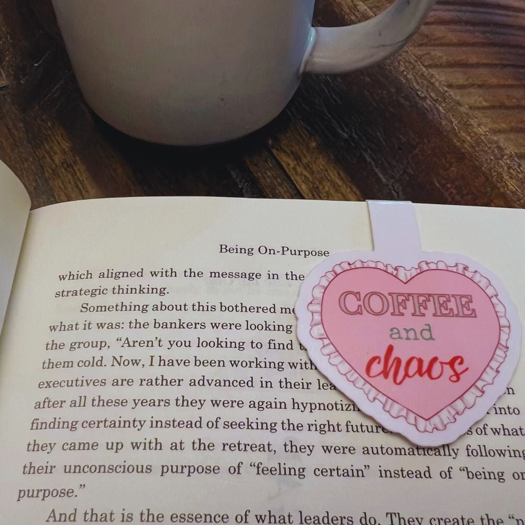 Pink Coffee & Chaos heart magnetic bookmark clipped over an open book beside a coffee mug for a cozy reader accessory.