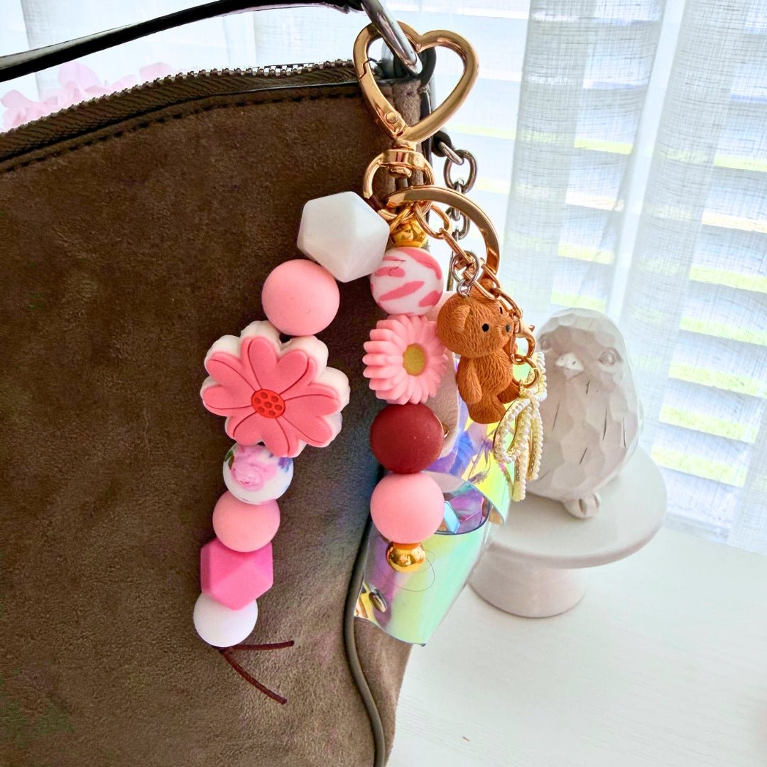 Finished pink silicone bead bag charm with flower accents, teddy bear charm, and holographic mini pouch attached to a handbag.