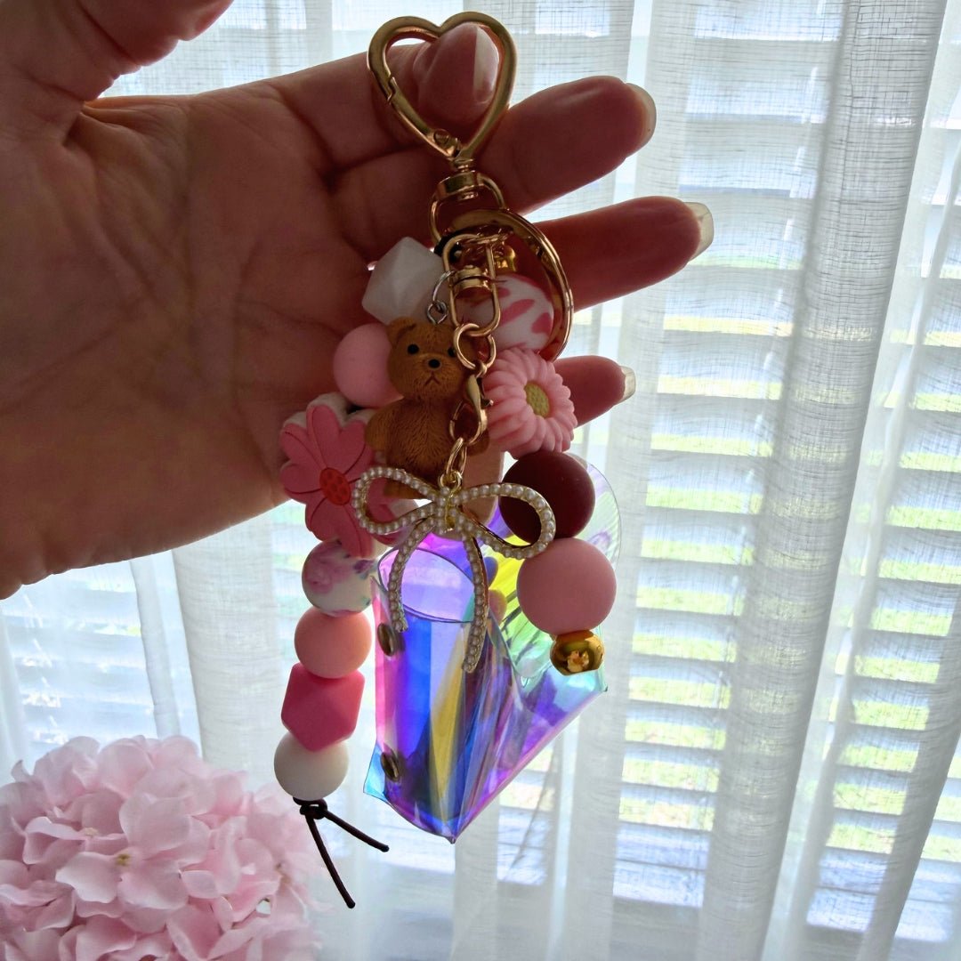 Hand holding a completed DIY bag charm with pink and white silicone beads, floral accents, gold clasp, and iridescent accessory pouch.