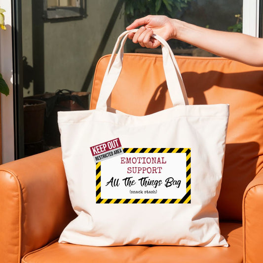 Canvas tote bag with “Emotional Support All The Things Bag” design held in hand on outdoor chair