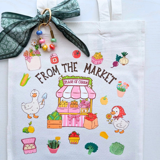 A white canvas tote bag with a colorful cartoon market illustration and the text 'FROM THE MARKET' and 'SPLASH OF CHARM'. The bag is styled with a green patterned fabric bow, beaded keychain, and small produce-themed accessories on a white background.