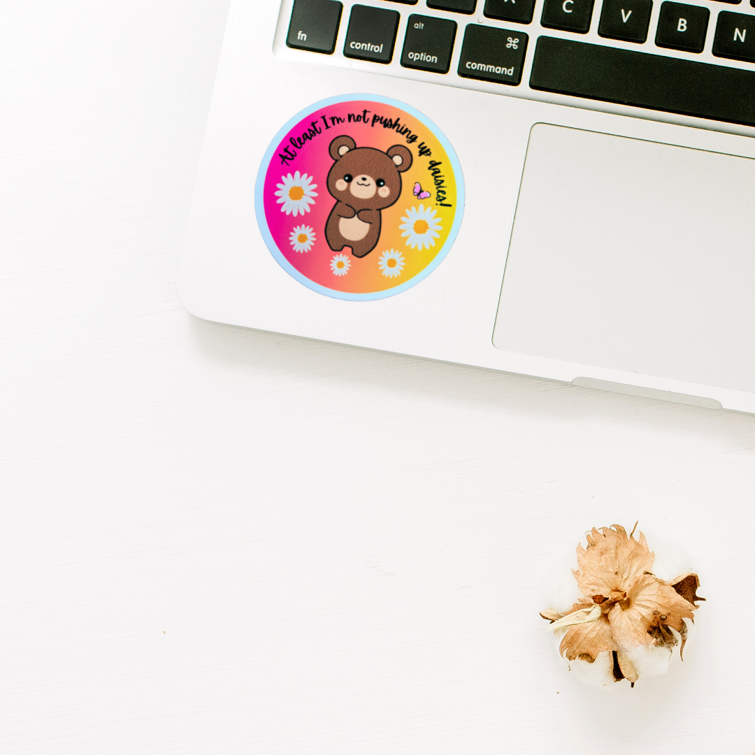 Funny bear sticker with “At Least I’m Not Pushing Up Daisies” text on colorful background placed on laptop
