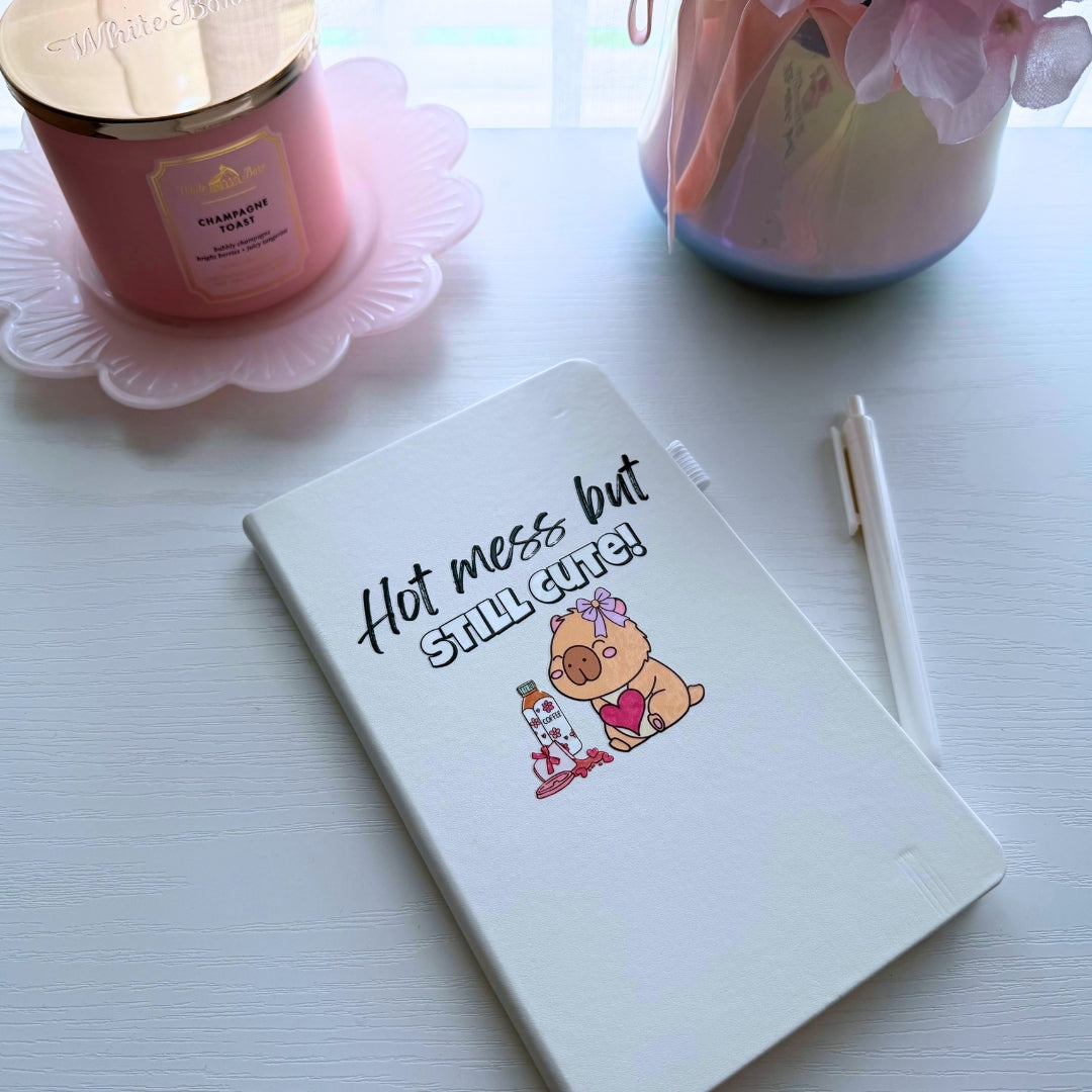 Hot Mess But Still Cute A5 hardcover journal displayed beside a candle, flowers, and pen on a cozy desk.