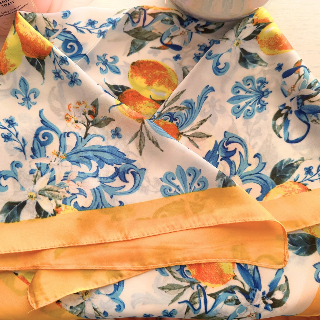 Square satin-feel polyester scarf featuring lemons, blue floral accents, and a yellow border displayed folded to show the vibrant spring-inspired print.