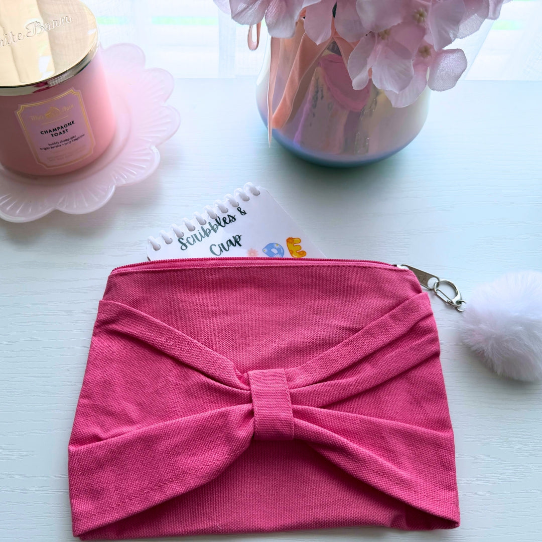 Pink canvas Mama zipper pouch with oversized bow and pom-pom zipper pull displayed with a notebook and cozy desk accessories.