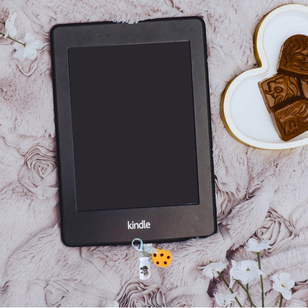 Cookies and milk dust cover charm attached to a Kindle on a pink textured blanket beside a heart-shaped dish of chocolates.