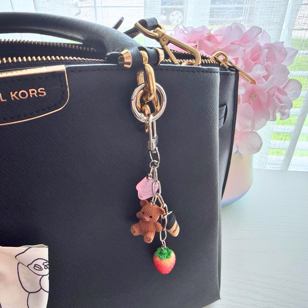 Strawberry teddy bear purse charm clipped to a black handbag featuring a coffee cup charm, pink star accent, and strawberry charm.
