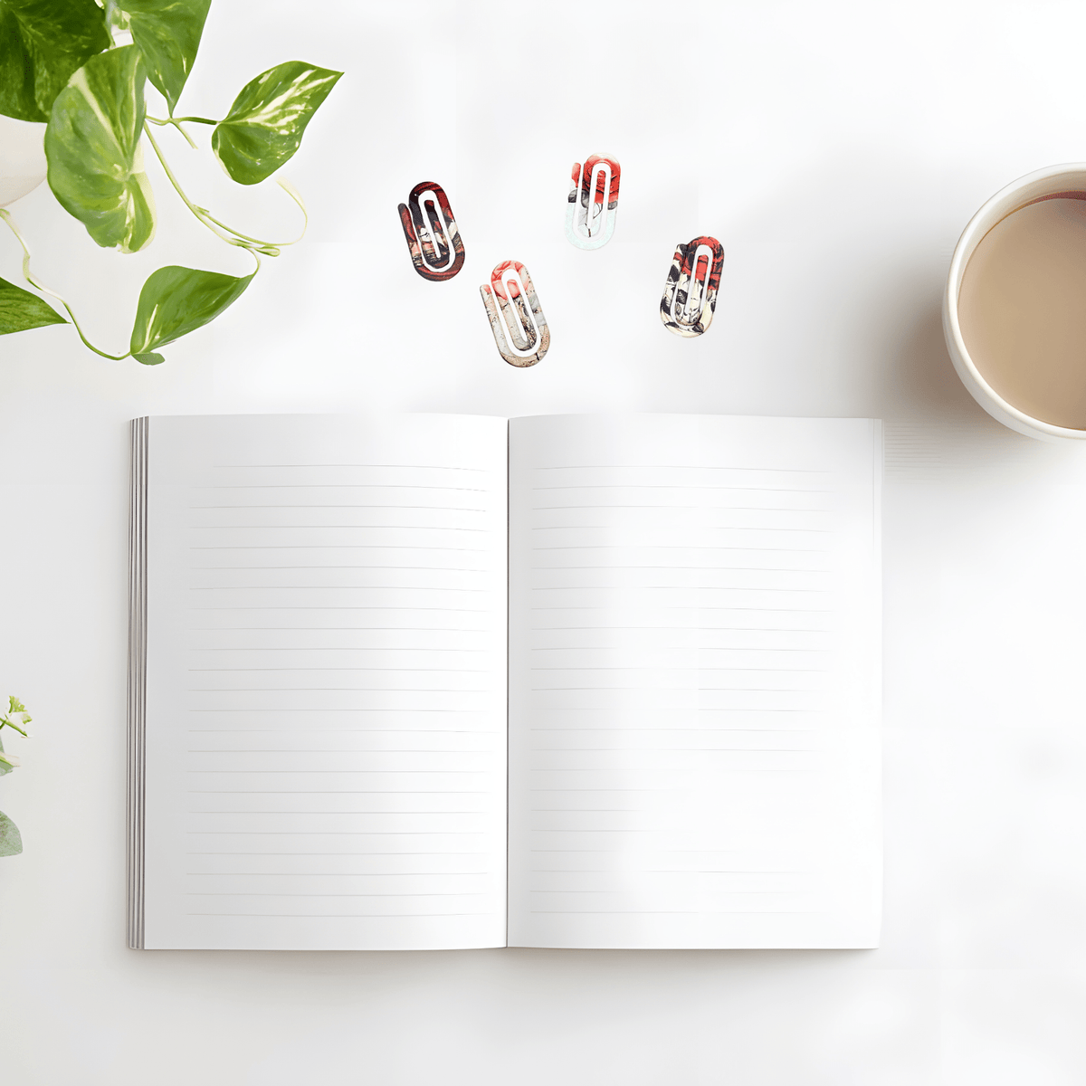 Open notebook with vintage-inspired paper clips on a white surface next to a cup of coffee and plant leaves.